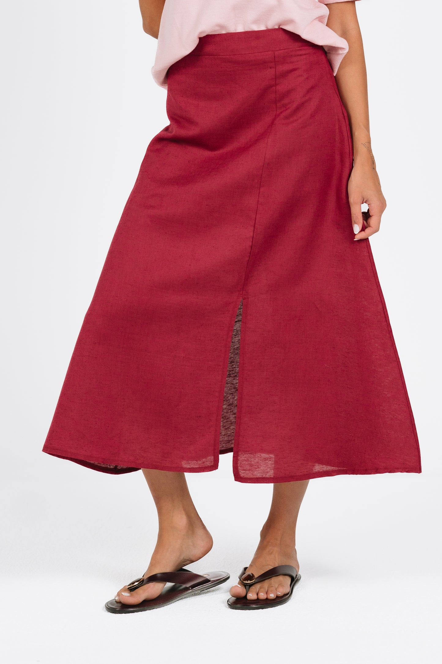 A woman wearing a high-waisted, midi-length red skirt with a side slit, styled with a light pink top and black flip-flop sandals against a plain white studio background.