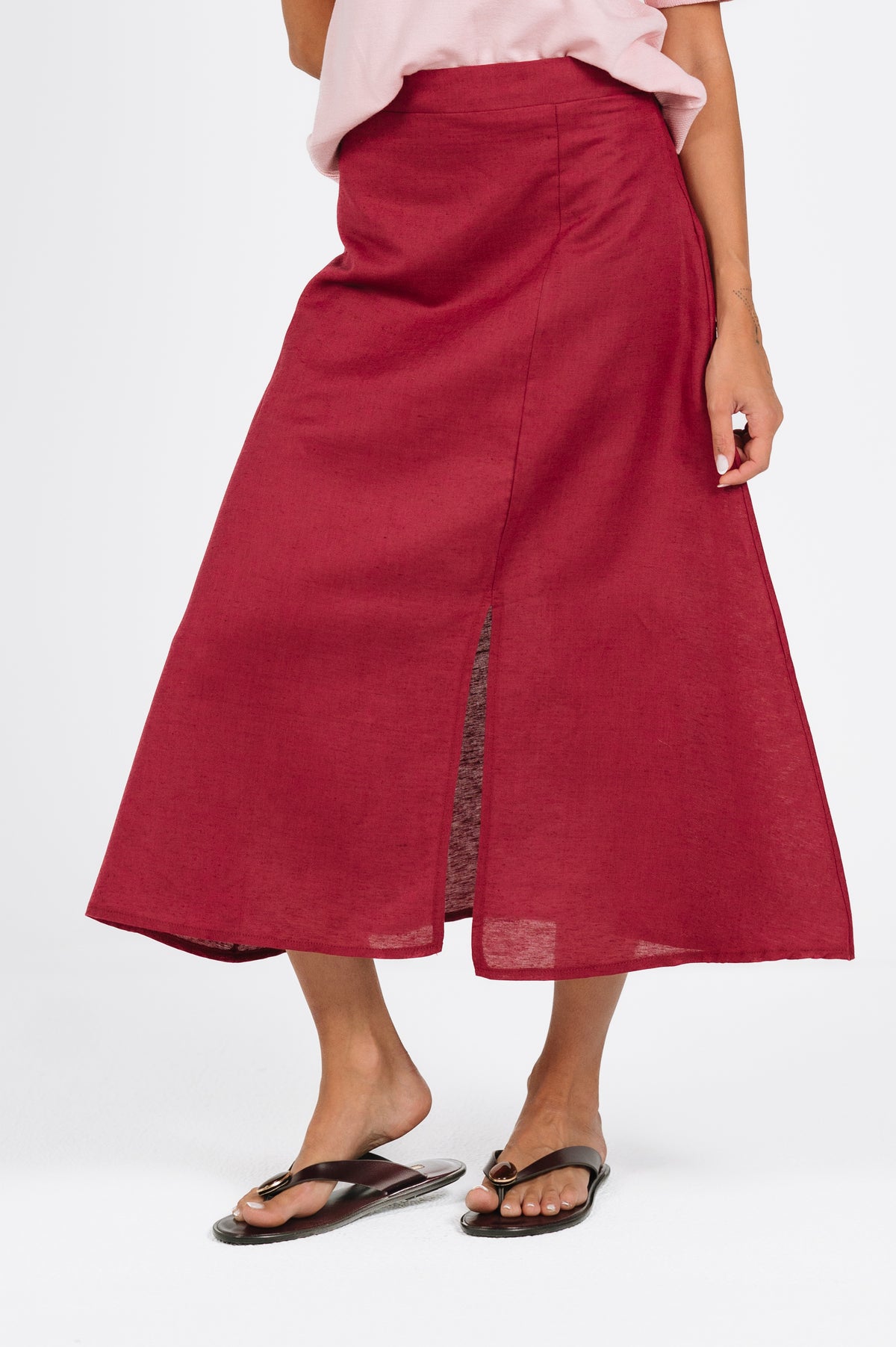 A woman wearing a high-waisted, midi-length red skirt with a side slit, styled with a light pink top and black flip-flop sandals against a plain white studio background.