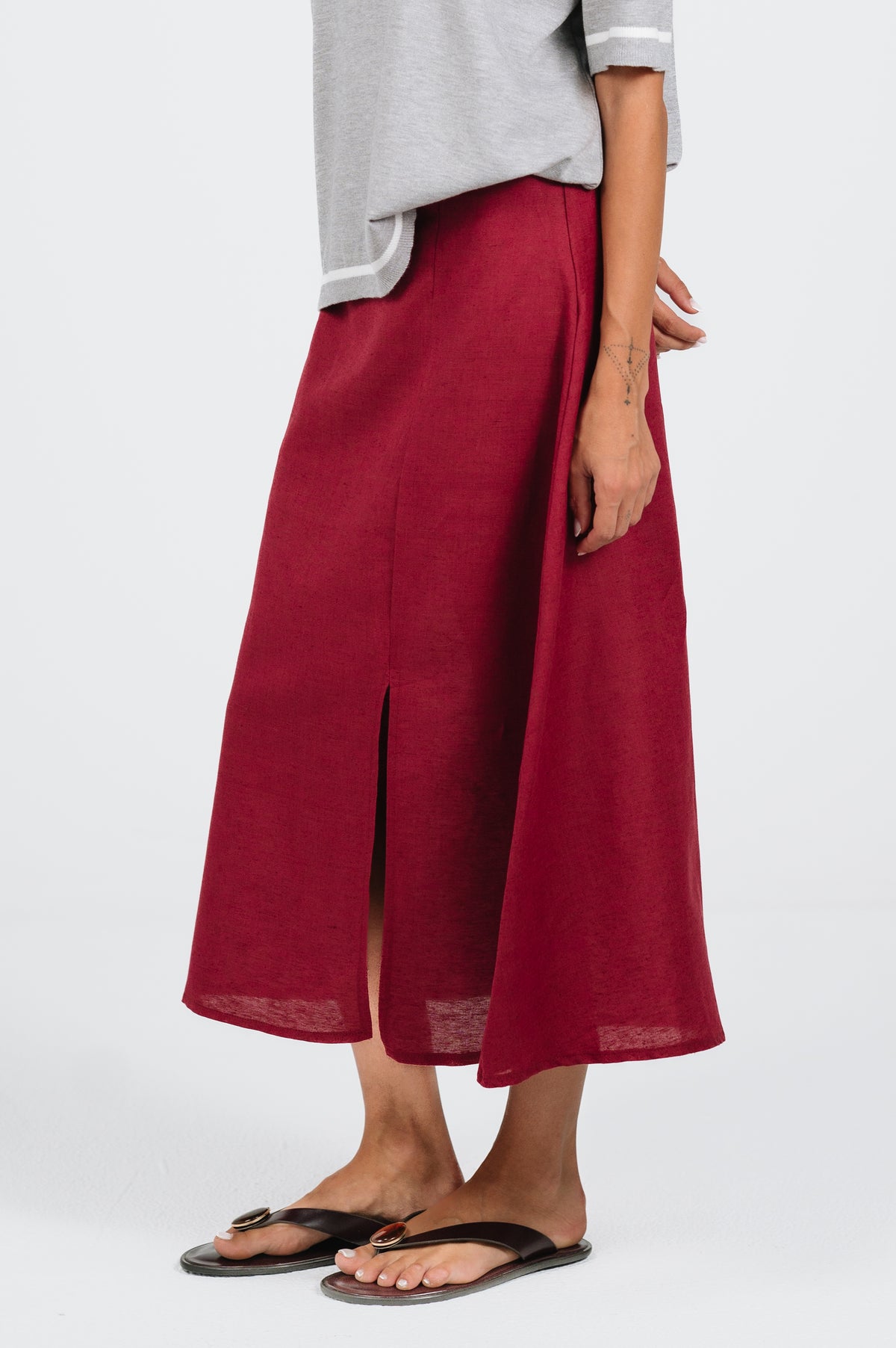 A person wearing a long, A-line burgundy skirt with a front slit, styled with a light gray top and black flip-flop sandals against a plain light gray background.